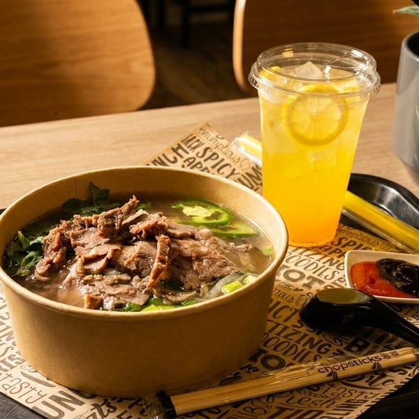 Beef pho in a paper bowl with a lemon drink, sauces, and chopsticks on a tray.